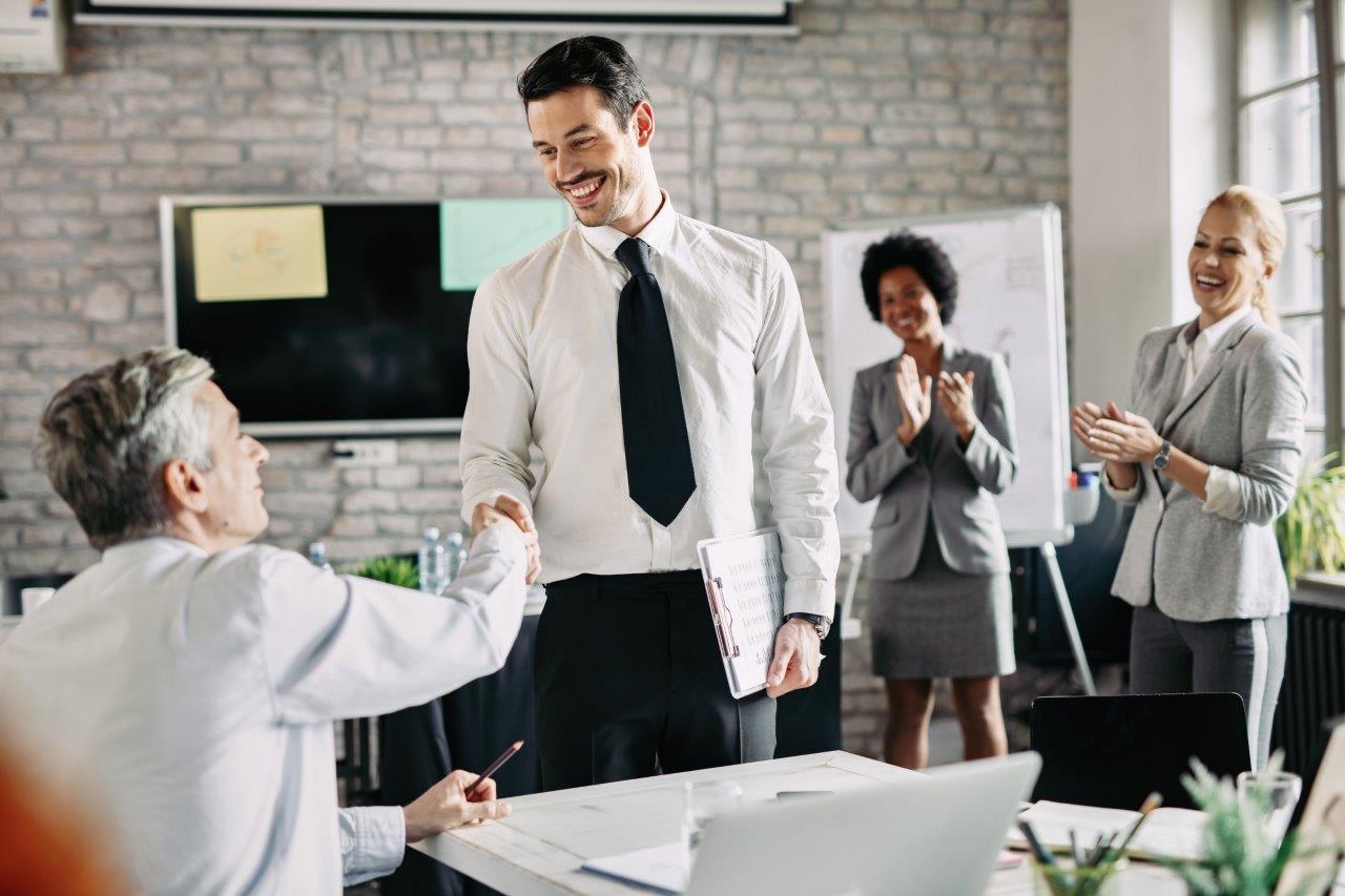 Two happy businessmen shaking hands after successful agreement office while their female colleagues are applauding them focus is young businessman